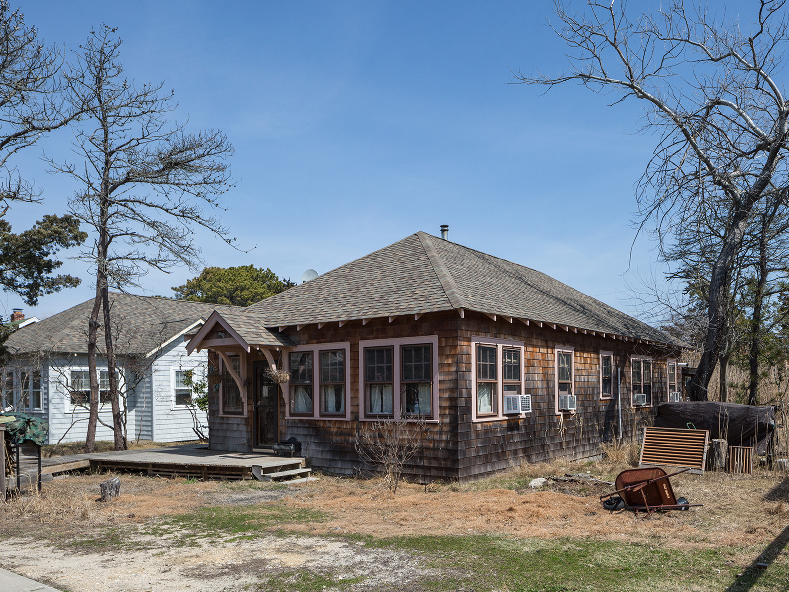 A wood-shingled bungalow with a small window sits on the ground on a sandy street is viewed from the southeast corner.