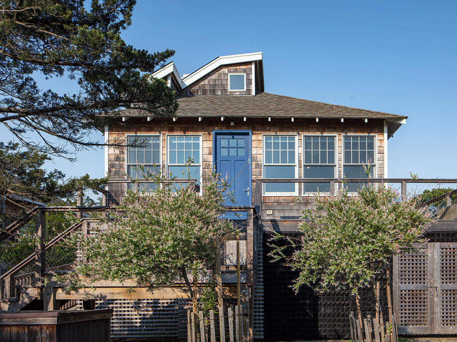 Front view of the wood-shingled house raised above flood level with young trees and a blue door