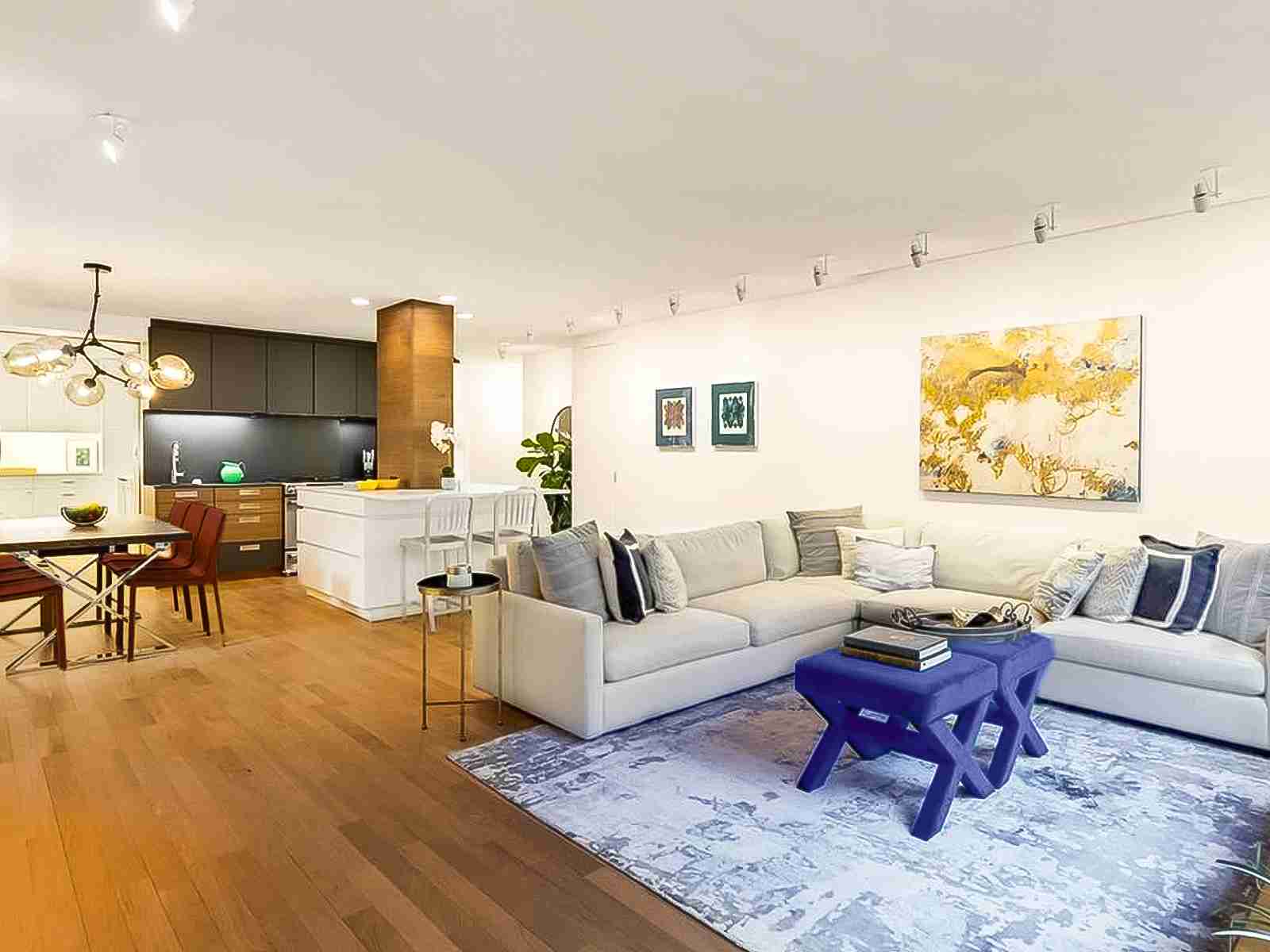 A large open plan Living Room opens to a dining table, a large white kitchen island with charcoal and walnut cabinetry beyond, and in the distance, a large pantry on the left and a corridor to the right