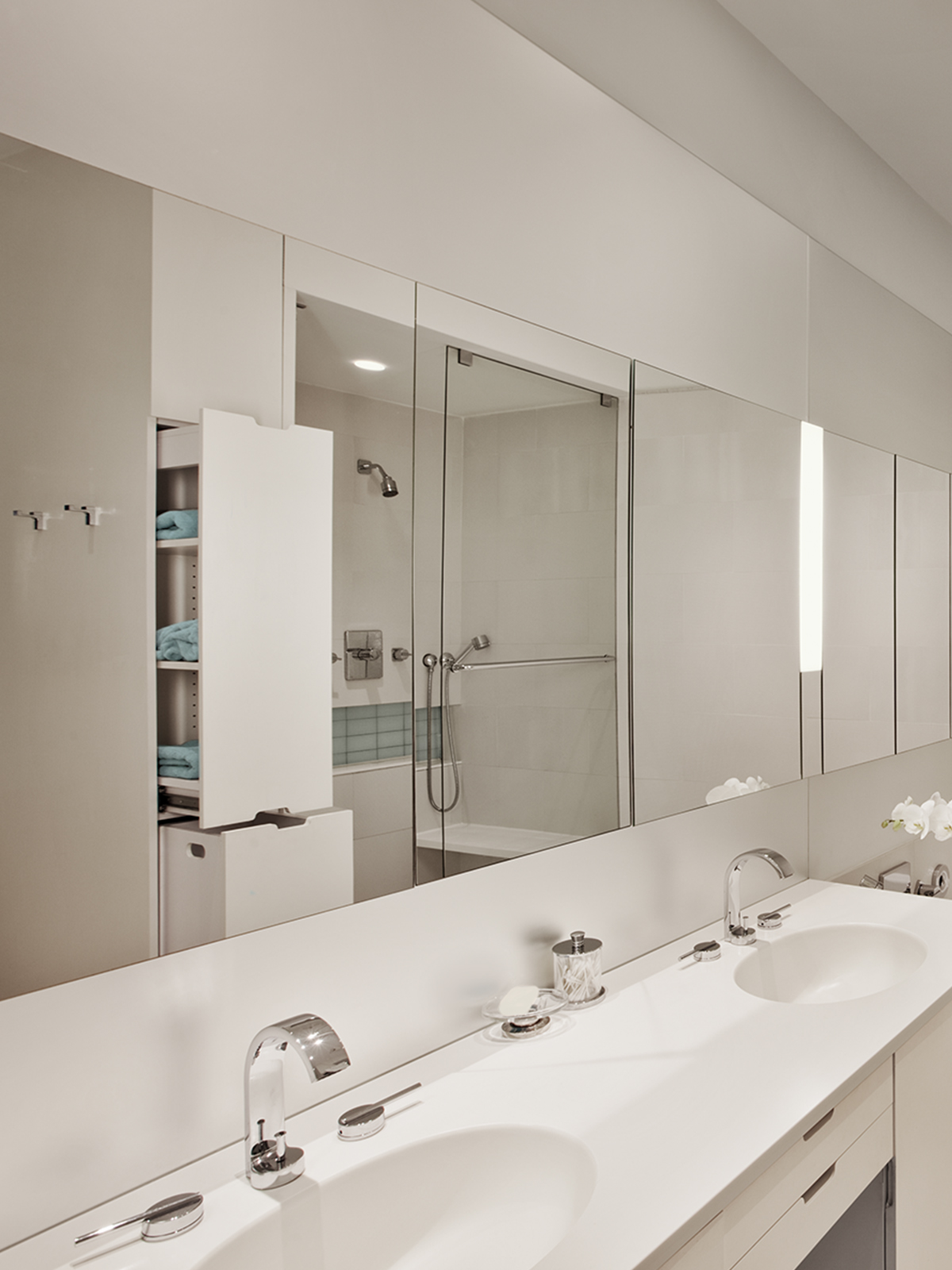 A glowing white bathroom with a double sink on the left, a double shower and pullout storage for towels and toiletries reflected in the mirror above the sinks, a tube in the center on the back wall of the bathroom