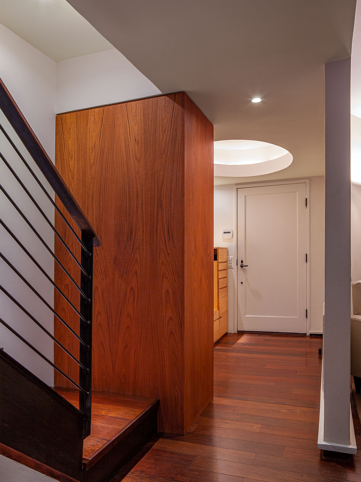 A teak paneled wall A divides the minimalist stair from the Entry. The low ceiling at the Entry is made higher with a recessed oval architectural light; wide wood floorboards connect the spaces.