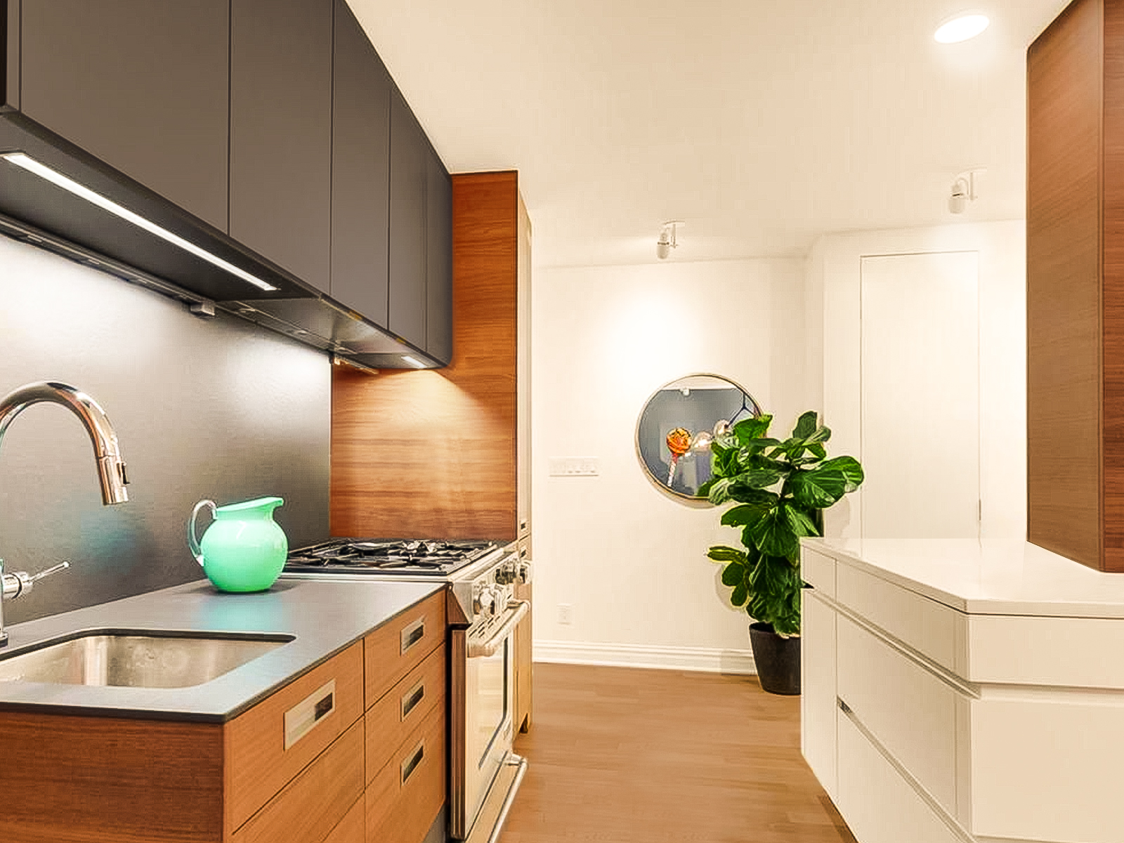 Looking toward the entry corridor, charcoal upper cabinets, charcoal countertops, and two-tone walnut drawers with flush stainless hardware hug the wall, the large white island anchored by a walnut clad column, is on the right