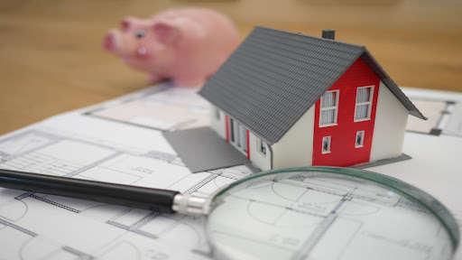 Wood tabletop with a house plan, toy house, a pink piggy bank, and a magnifying glass.