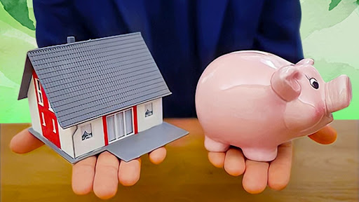 two hands hold a pink piggy bank on the right, and a toy house on the left, over a wood table-top against a leafy green background.
