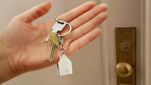 A hand holds keys on a keychain with a small house charm in front of a white, wooden door.