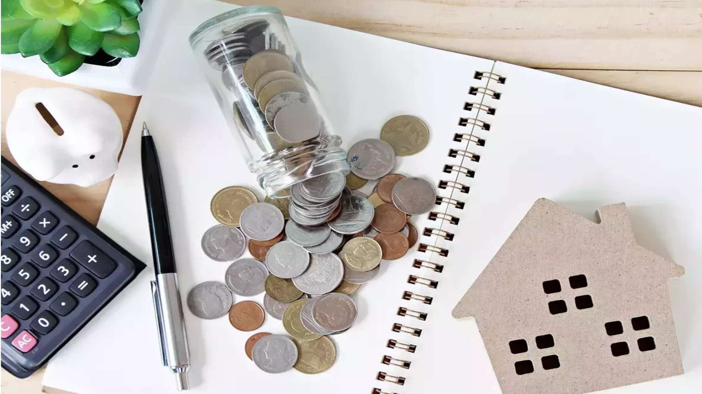 Notebook on a table with a calculator, a piggy bank, scattered coins, a pen, and a cardboard cutout of a house.