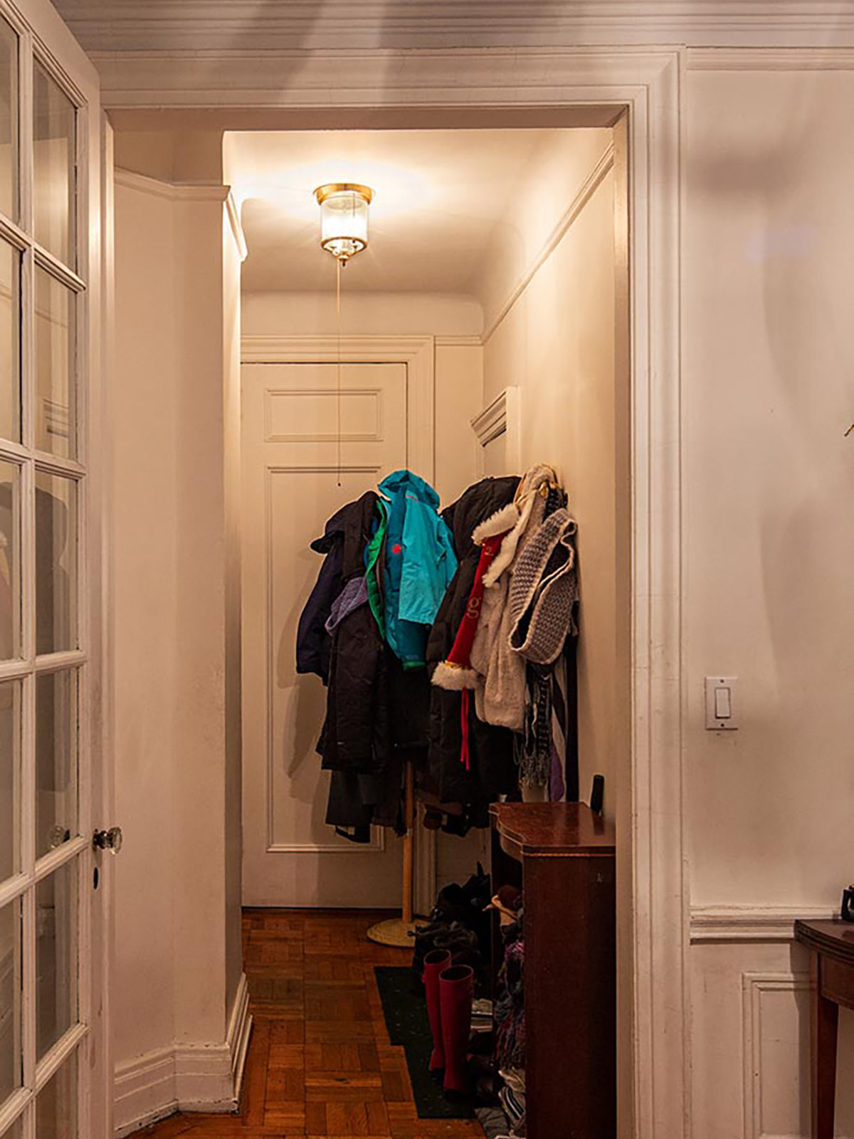 Entry vestibule in a prewar apartment crammed with coat racks that block the small closet, the paint is old, and the floor is worn.