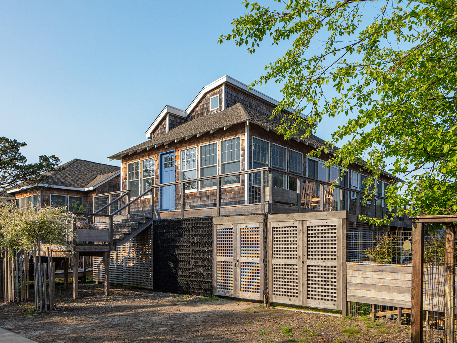 Transformed wood-shingled bungalow with wall to wall-to-wall windows raised above flood level seen from its southeast corner.