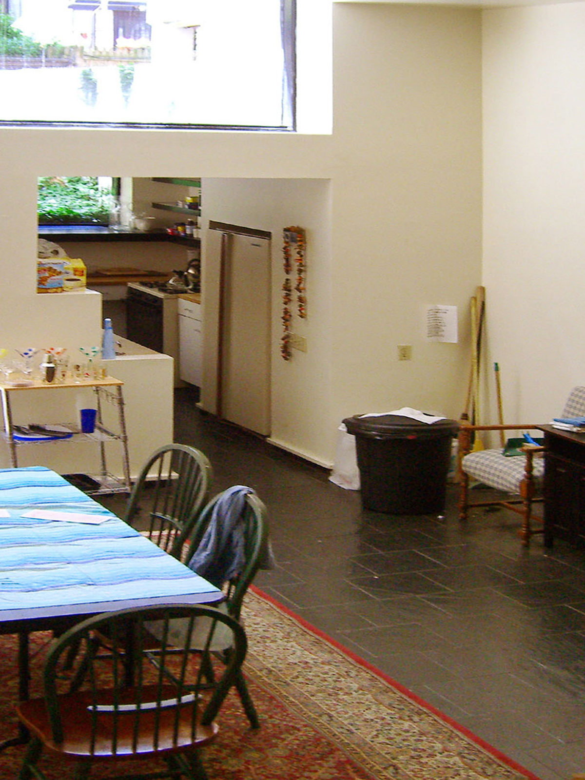 Looking down on double-height dining space, a table with a blue plastic covering, and empty walls. A stepped sheetrock divider obscures a Galley Kitchen