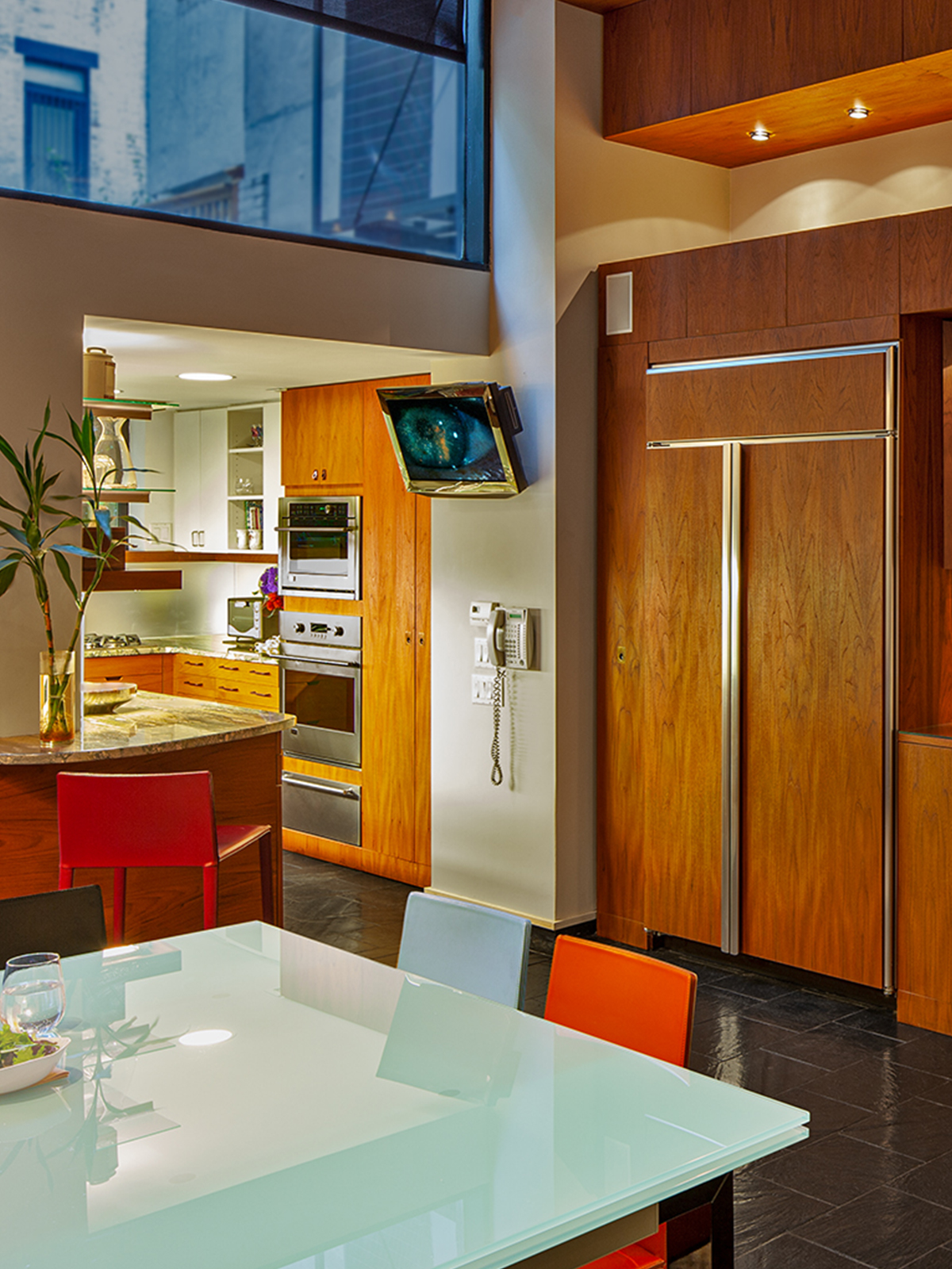 (The same view) A glass dining table gleams in the foreground, a stool at a stone countertop wraps the corner into the Galley Kitchen, and flat teak paneled refrigerator continue the cabinetry from the Kitchen into the Dining Room