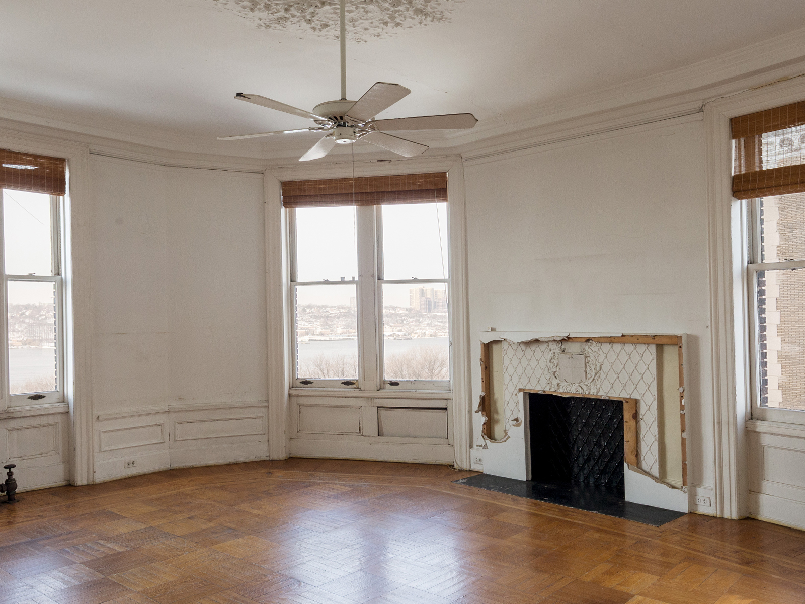 An unrestored room with large windows and fireplace surround revealed behind torn-open sheetrock