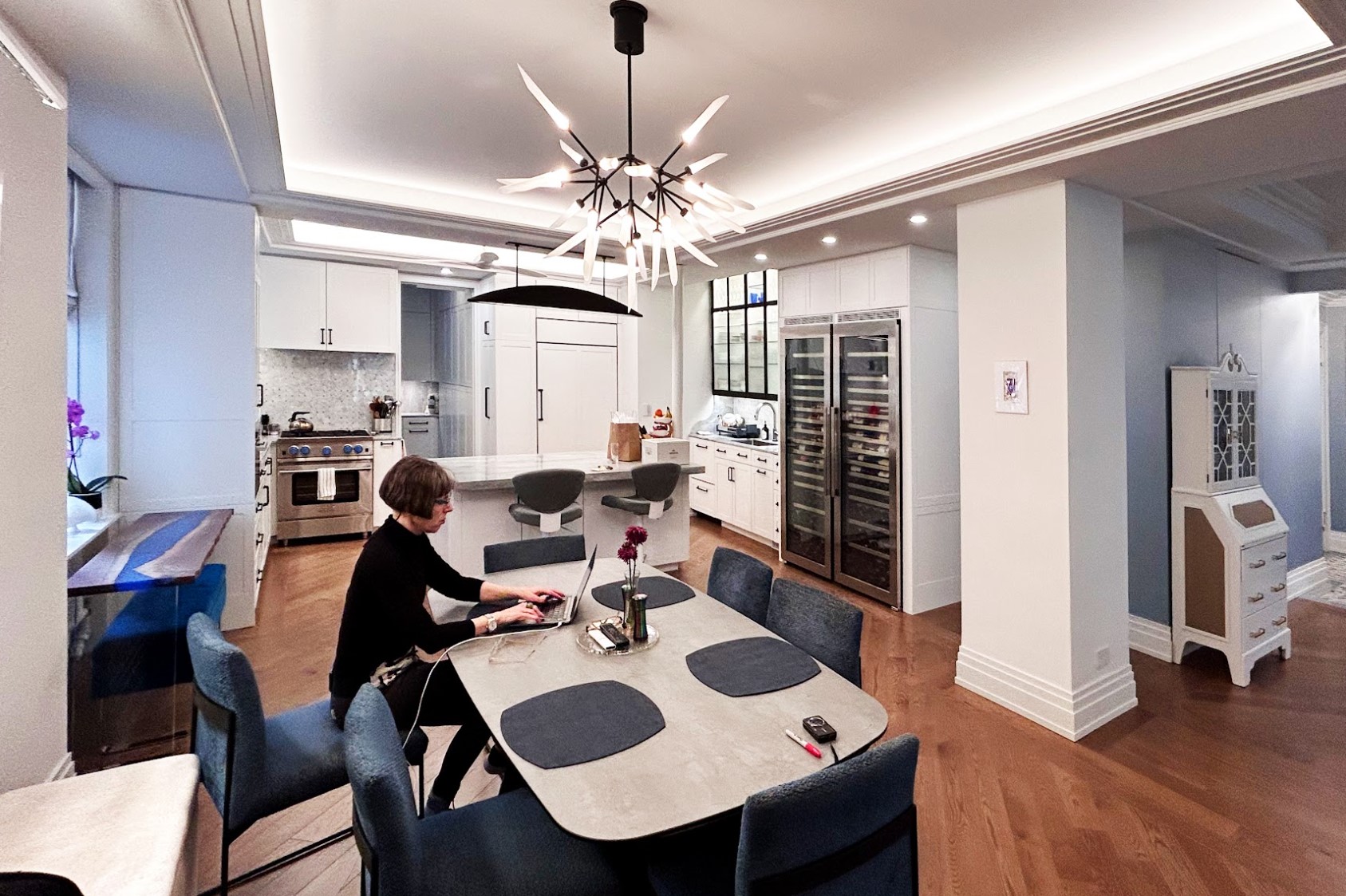 After: The corner is now a wide-angle view of a chef’s Kitchen with glass-fronted wine refrigerators and lighted custom metal cabinets display glassware to the right.