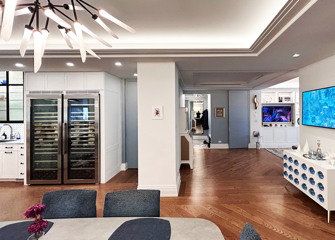 After: The renovated dining room is expansive. The dining table is in the foreground, and glass-fronted wine refrigerators and lighted custom metal cabinets display glassware in the background. The walls have been moved so that the daylight entering from the living room is visible.