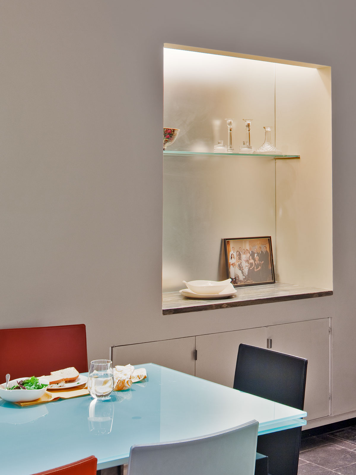 (the same view) Chairs of different colors surround a glass table; the niche now has architectural lighting and frosted mirror accent the niche