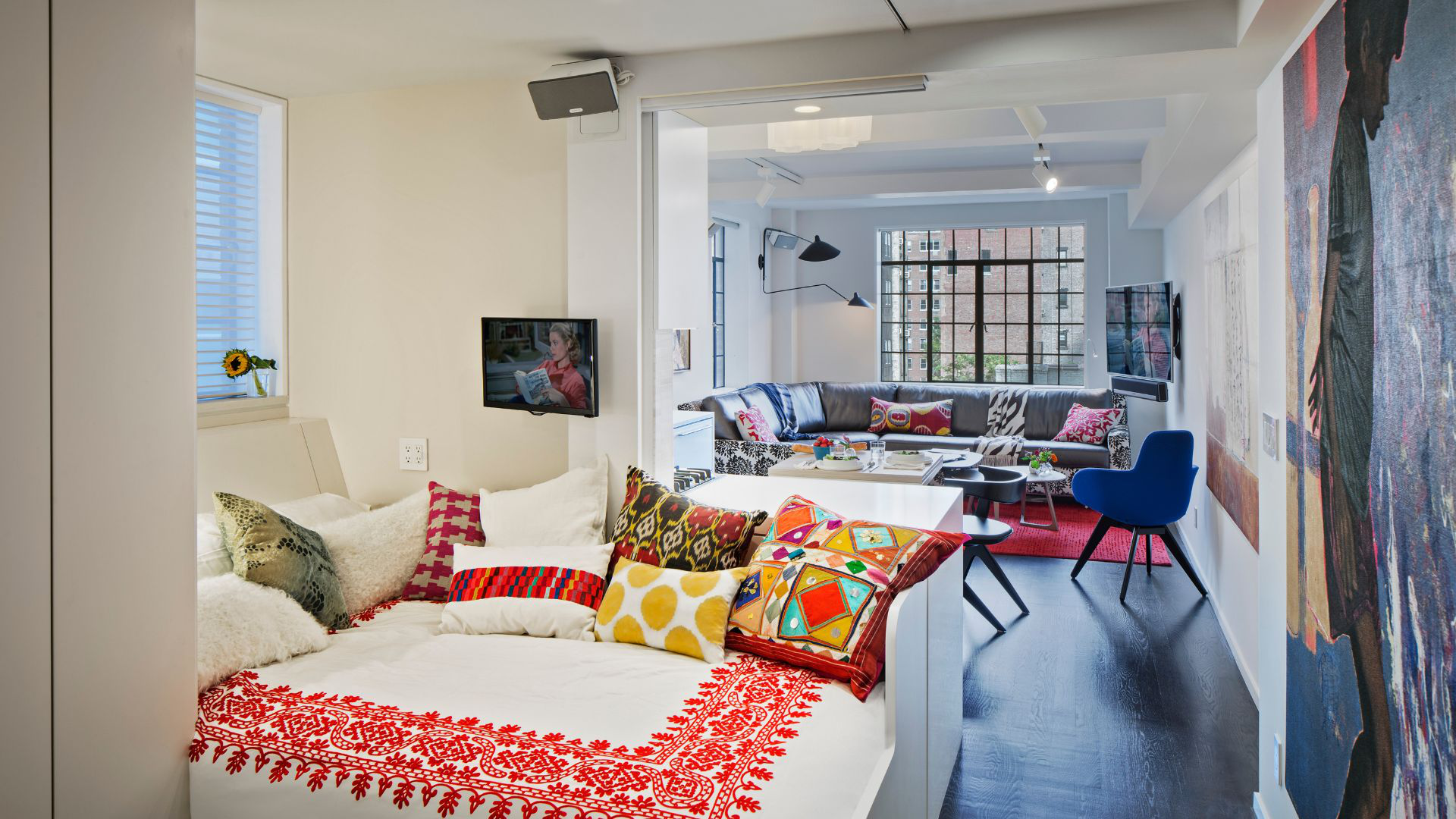 Dark floors set off a white bed adorned with vibrant, multicolored pillows and covered with a view through the apartment to the large window and living area beyond.