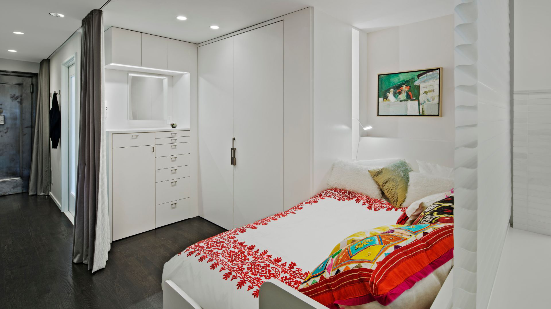 A cozy, curtained sleeping alcove white with red accents on the bedding. A small painting and light slot personalize the space. A built-in dresser and a wide closet are on the far side.