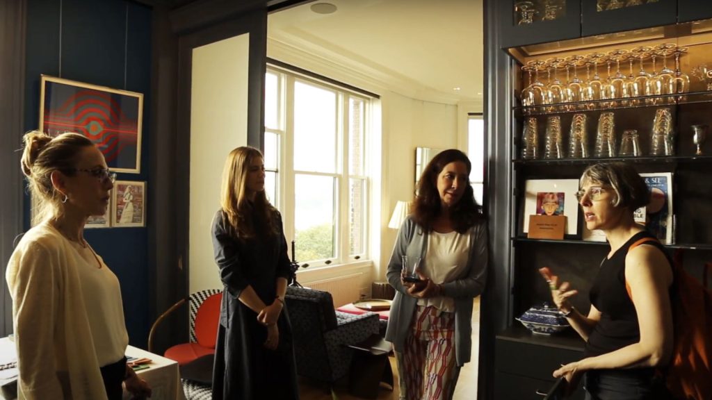 In a room with a large doorway, views of 2 windows, and architectural lighting illuminating dozens of glasses, four people admire the space.