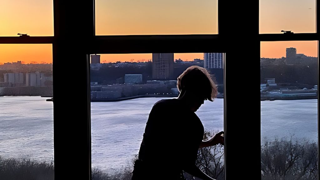 Allegra stands by a window that fills the frame. The warm glow of sunset sits on the horizon across the Hudson River.