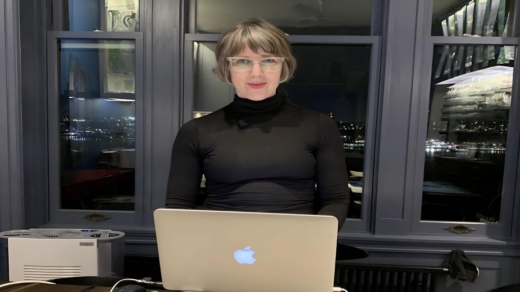 Allegra smiles at the photographer from behind an open laptop. The night-time reflection in the large window behind her mirrors her illuminated kitchen cabinets.