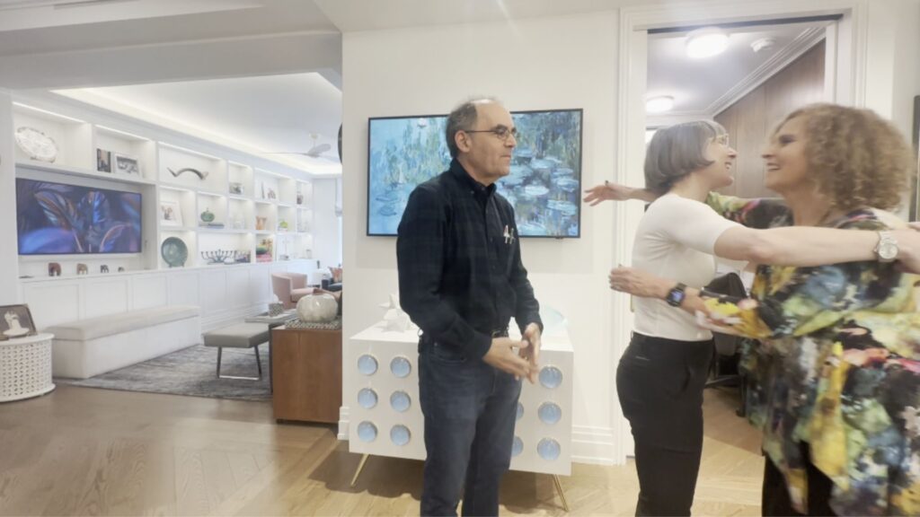 Allegra, the architect (center), engages with her clients, Steve (left) and Marcia (right), with Allegra and Marcia smiling and reaching out to hug each other in an open room with pale brown floors, white walls, and colorful art. Two rooms are visible beyond: on the left, a long open bookshelf with a TV screen and art objects extends into the distance along the living room, and on the right, a wood-paneled passageway.